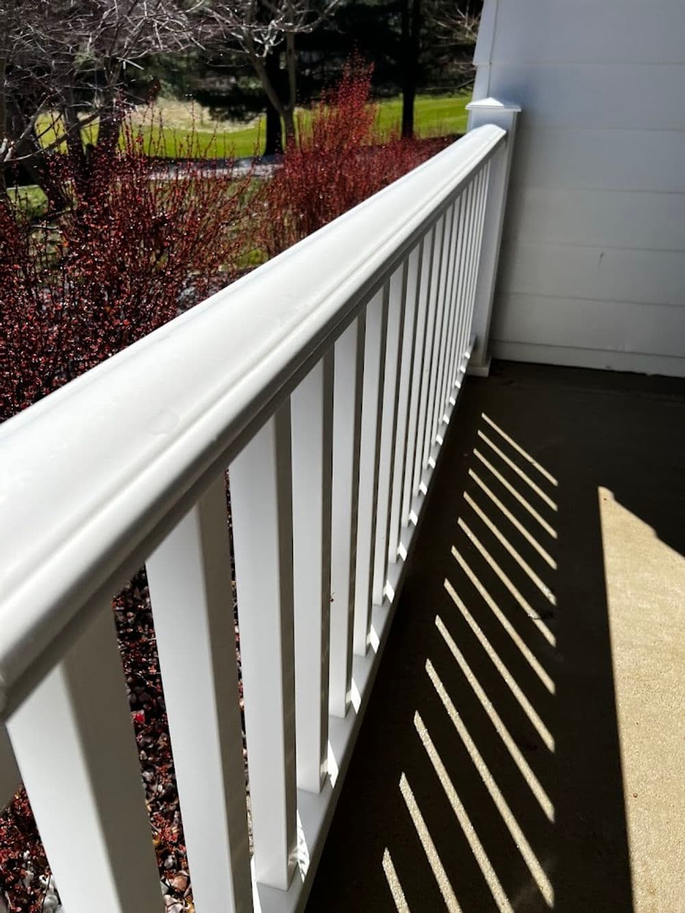 White railing casting shadows on a patio, with a backdrop of red foliage and greenery.