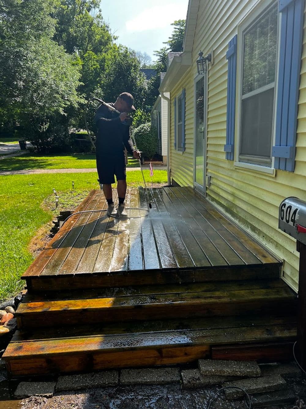Man pressure washing a wooden deck on a sunny day, surrounded by greenery and a yellow house.