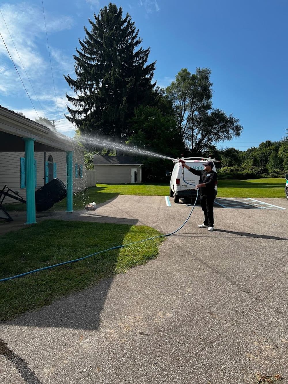 Person washing a building exterior with a hose on a sunny day, surrounded by greenery.