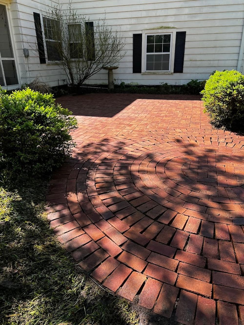 Brick patio with curved pattern and green shrubs against a white house backdrop.