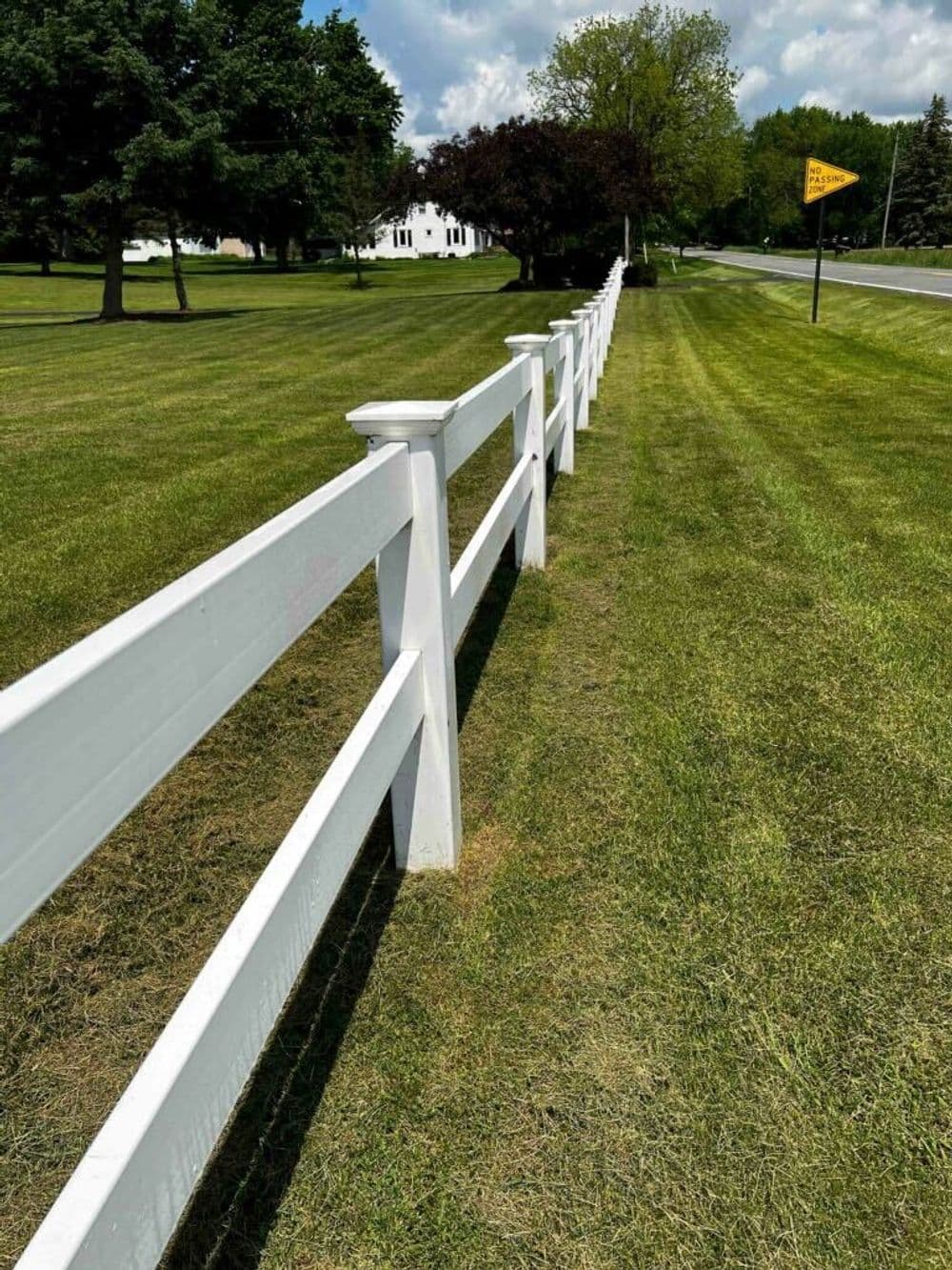 White wooden fence along a grassy lawn with trees and a road sign in the background.
