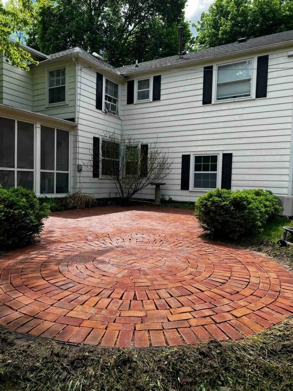 Brick patio design featuring a circular pattern, surrounded by greenery and a house exterior.