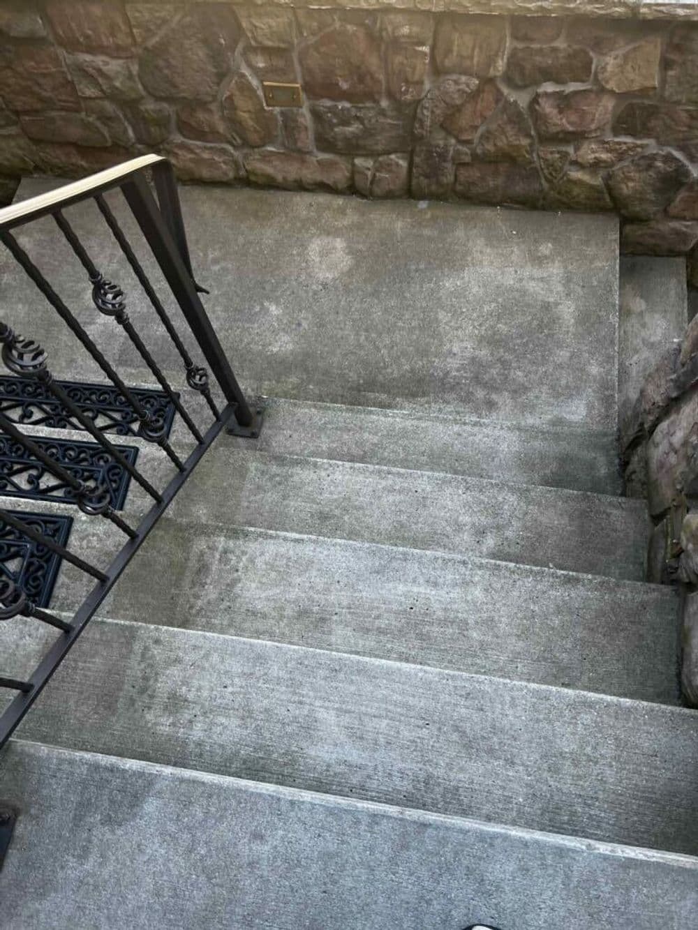 Concrete stairs with a black wrought iron railing and stone wall backdrop.