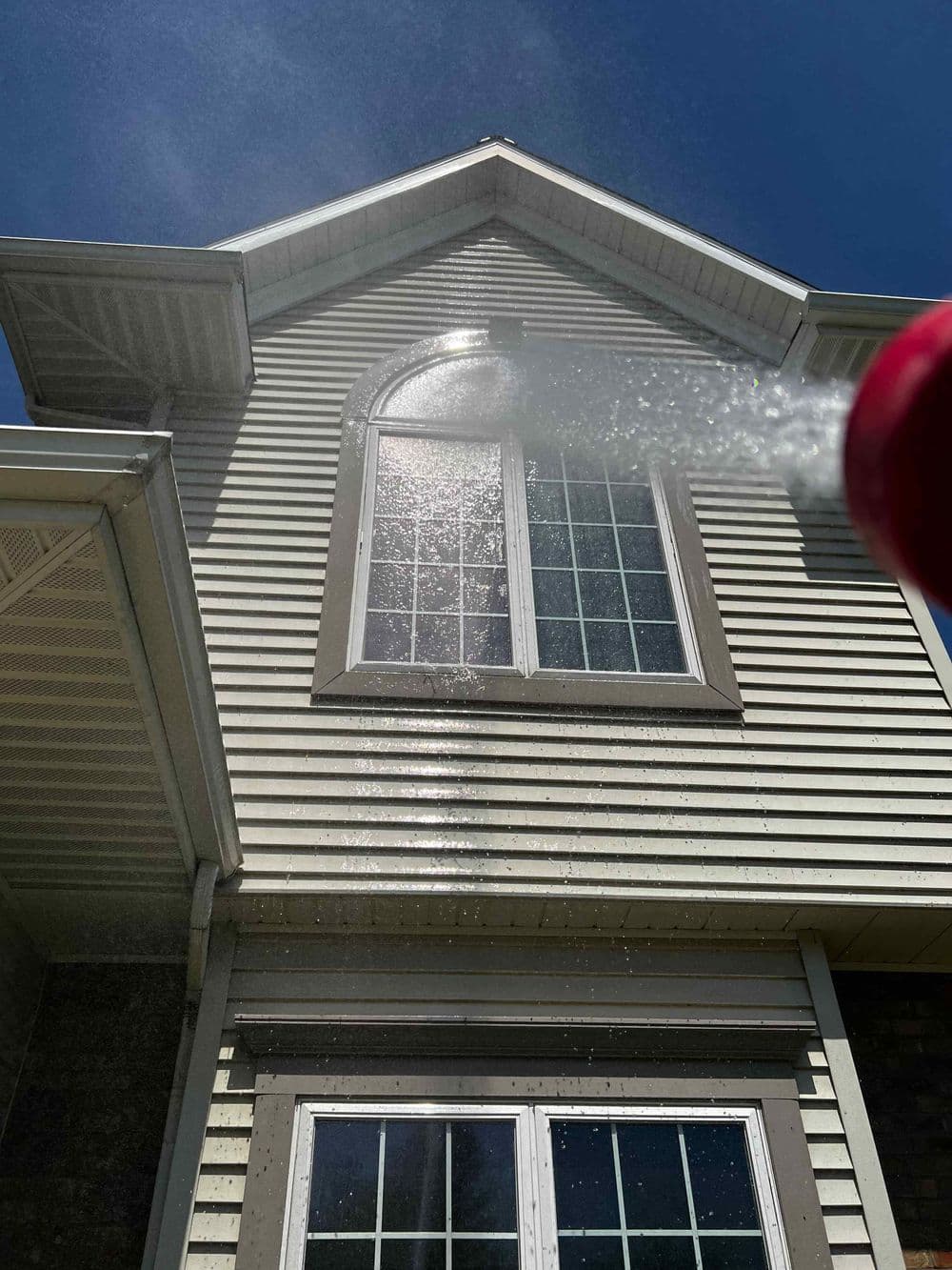Water spraying on a second-story window during home cleaning under bright sunlight.
